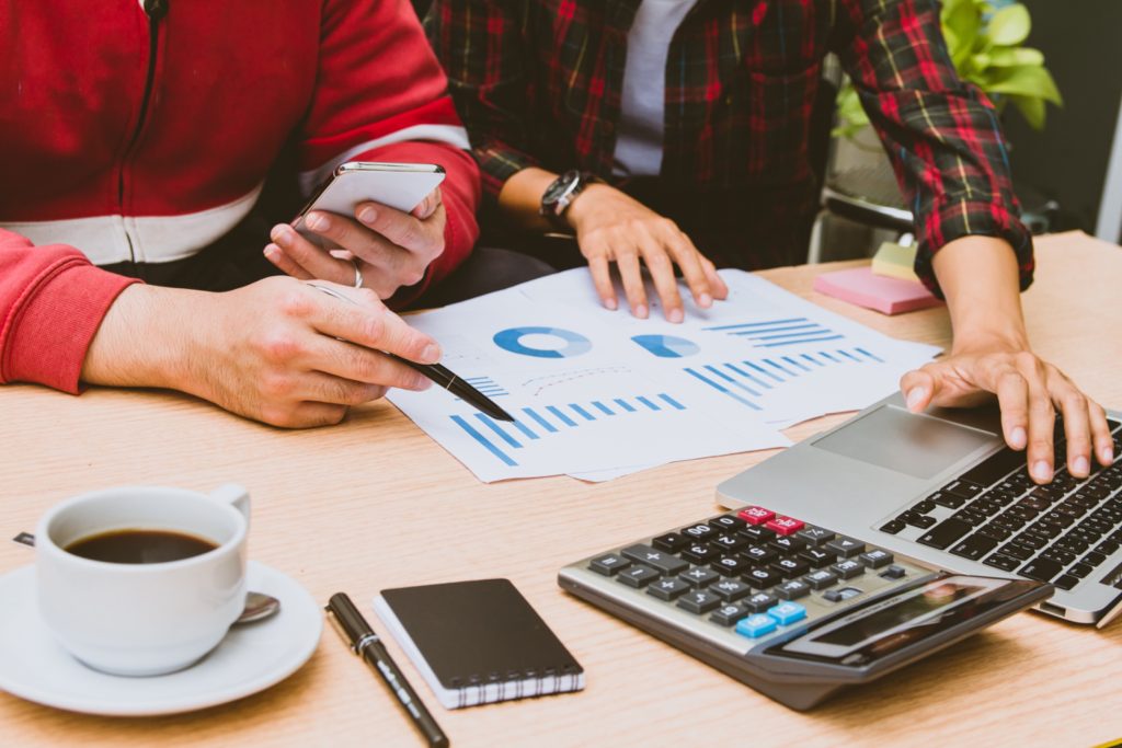 Hands reviewing financial charts beside a laptop, calculator, and coffee