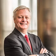 Professional headshot of a smiling businessman in dark suit and pink tie with arms crossed